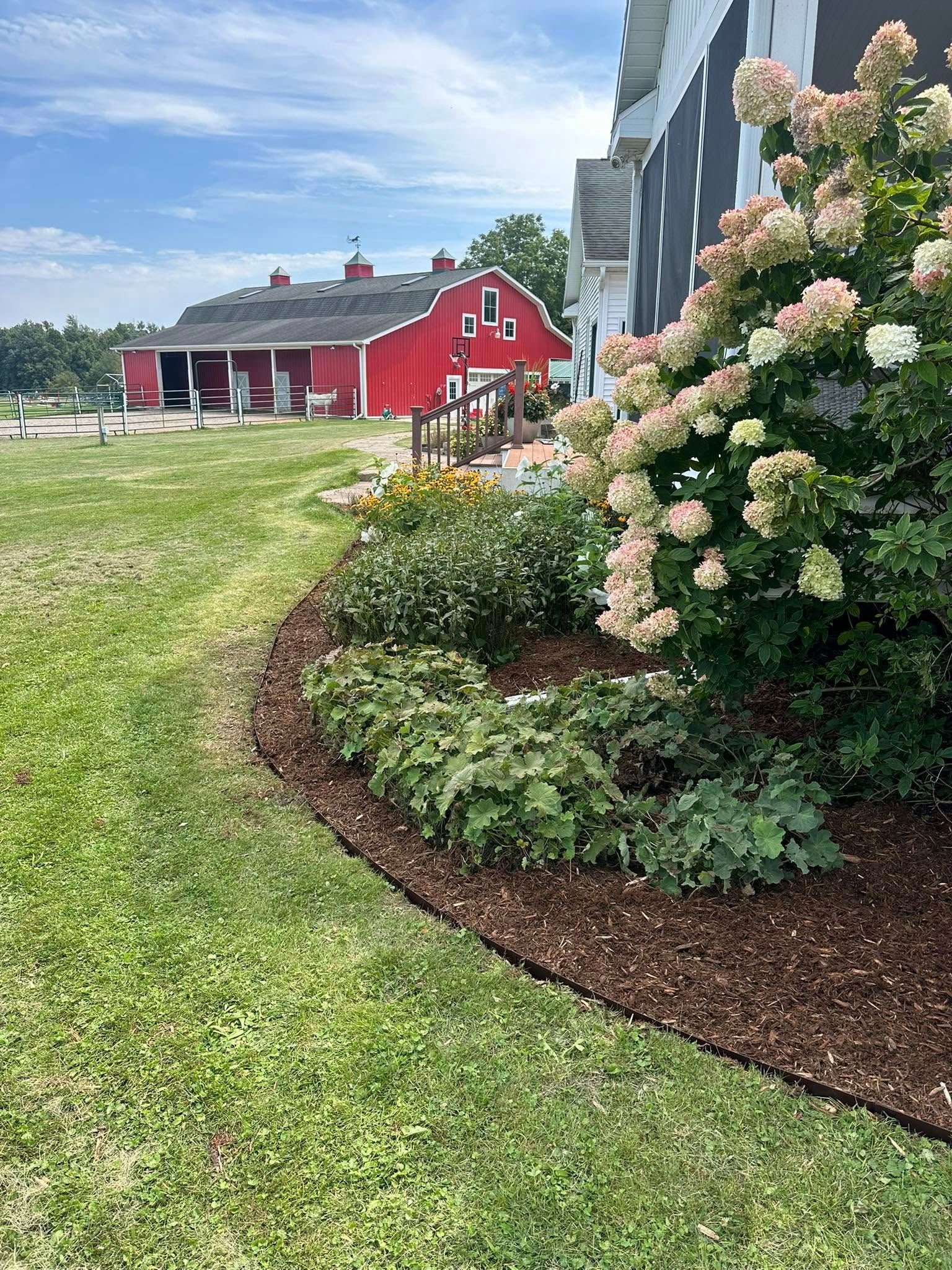 Red barn and garden bed with flowers and mulch, set on grassy area.