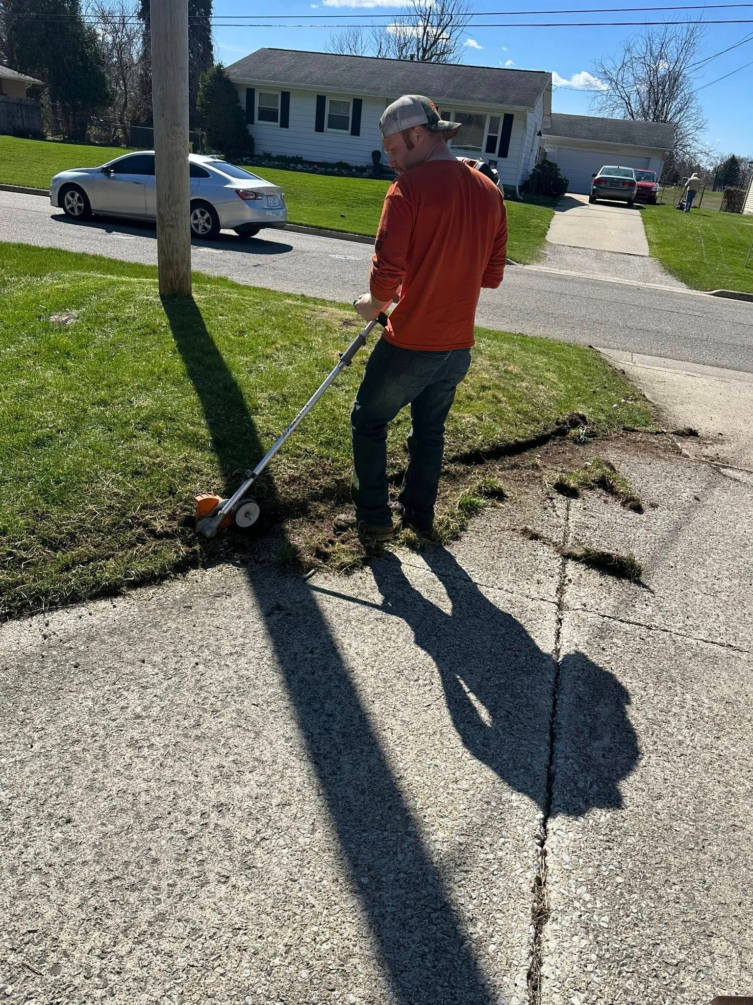Man in orange shirt using a weed whacker along a sidewalk near a grassy area with a utility pole.