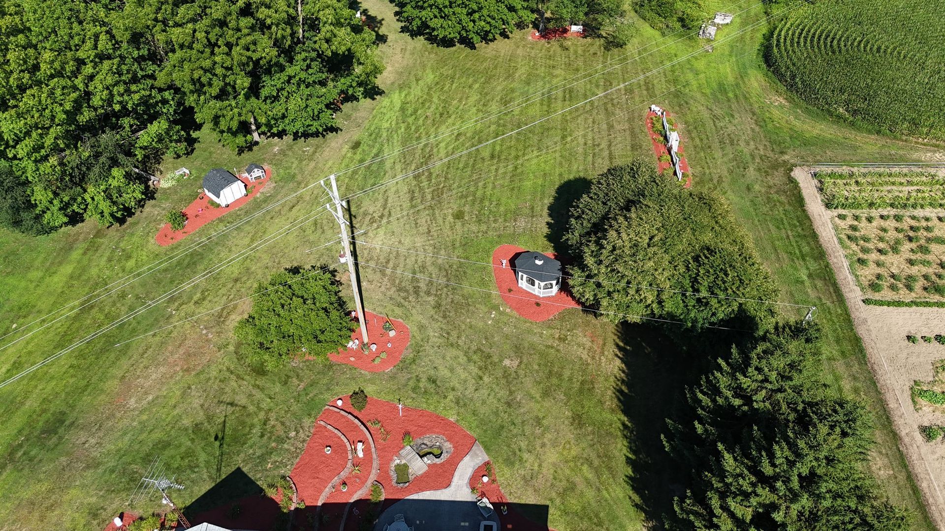 Aerial view of a grassy landscape with small buildings, gardens, trees, and power lines.