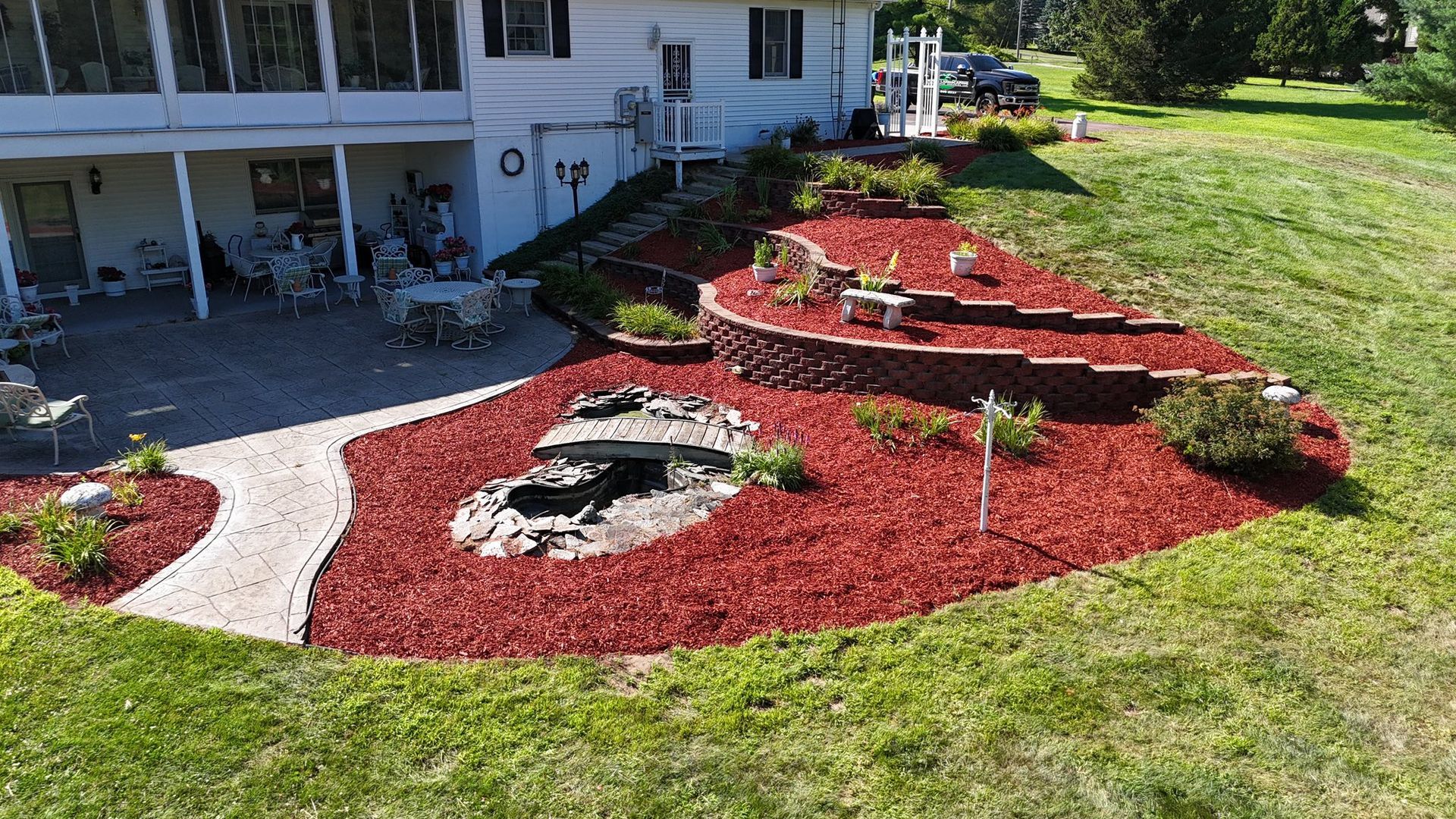 Lush green grass borders a bed of brown mulch with budding shrubs, next to a gravel path and trees.