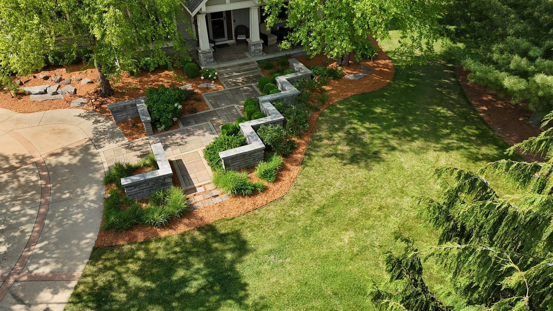 Lush green grass borders a bed of brown mulch with budding shrubs, next to a gravel path and trees.