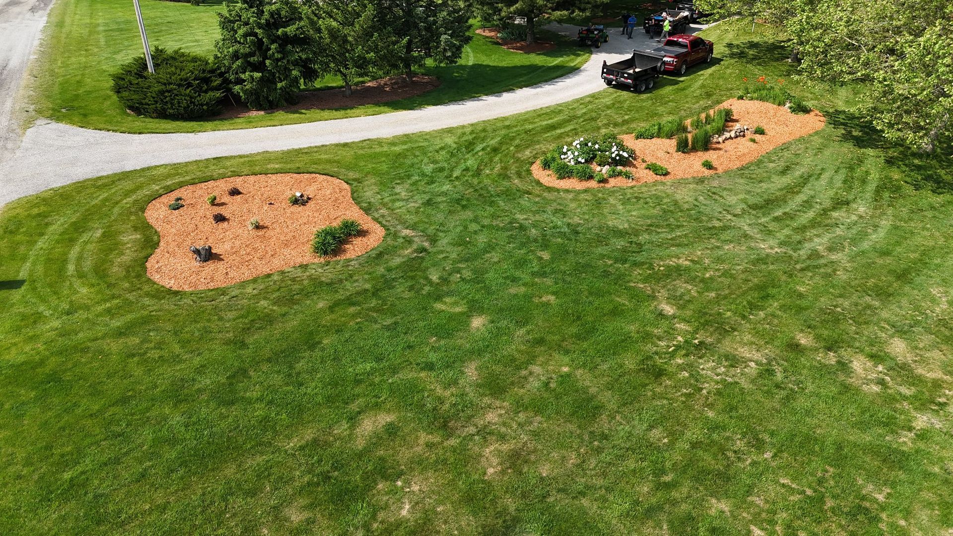 Green lawn with two mulch-covered flowerbeds and gravel driveway. A vehicle is parked in the distance.