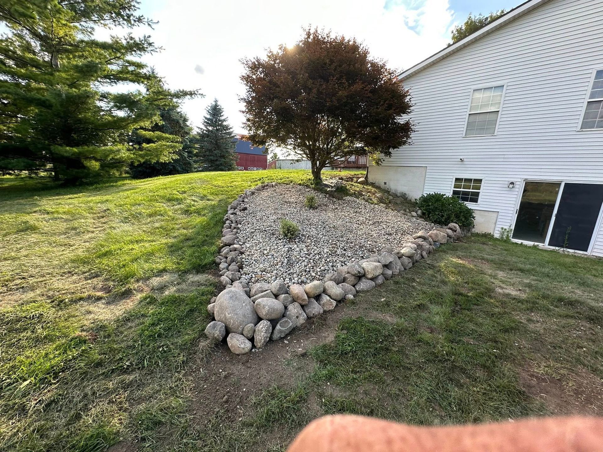 Rock-bordered flower bed on a slight hill by a house with a tree. Green grass and blue sky.