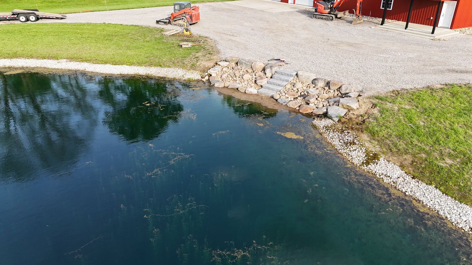 Pond with rocky shoreline and steps, next to a gravel area near red building and green grass.