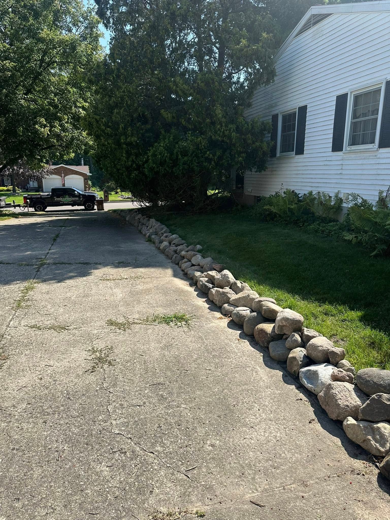 A driveway with a rock border next to a lawn and a white house. A trailer is in the background.