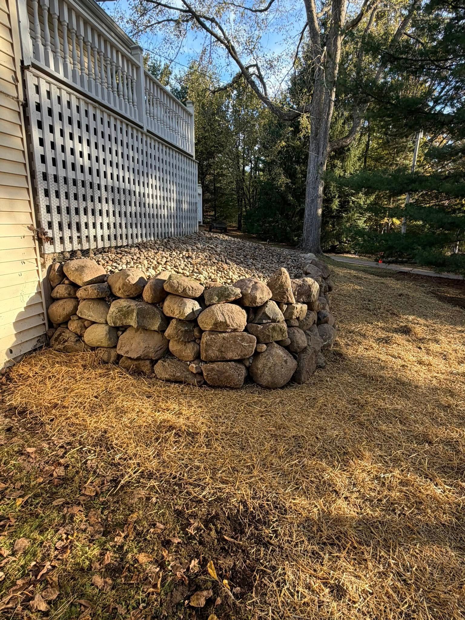 Stone retaining wall next to a building, surrounded by mulch, and trees in the background.