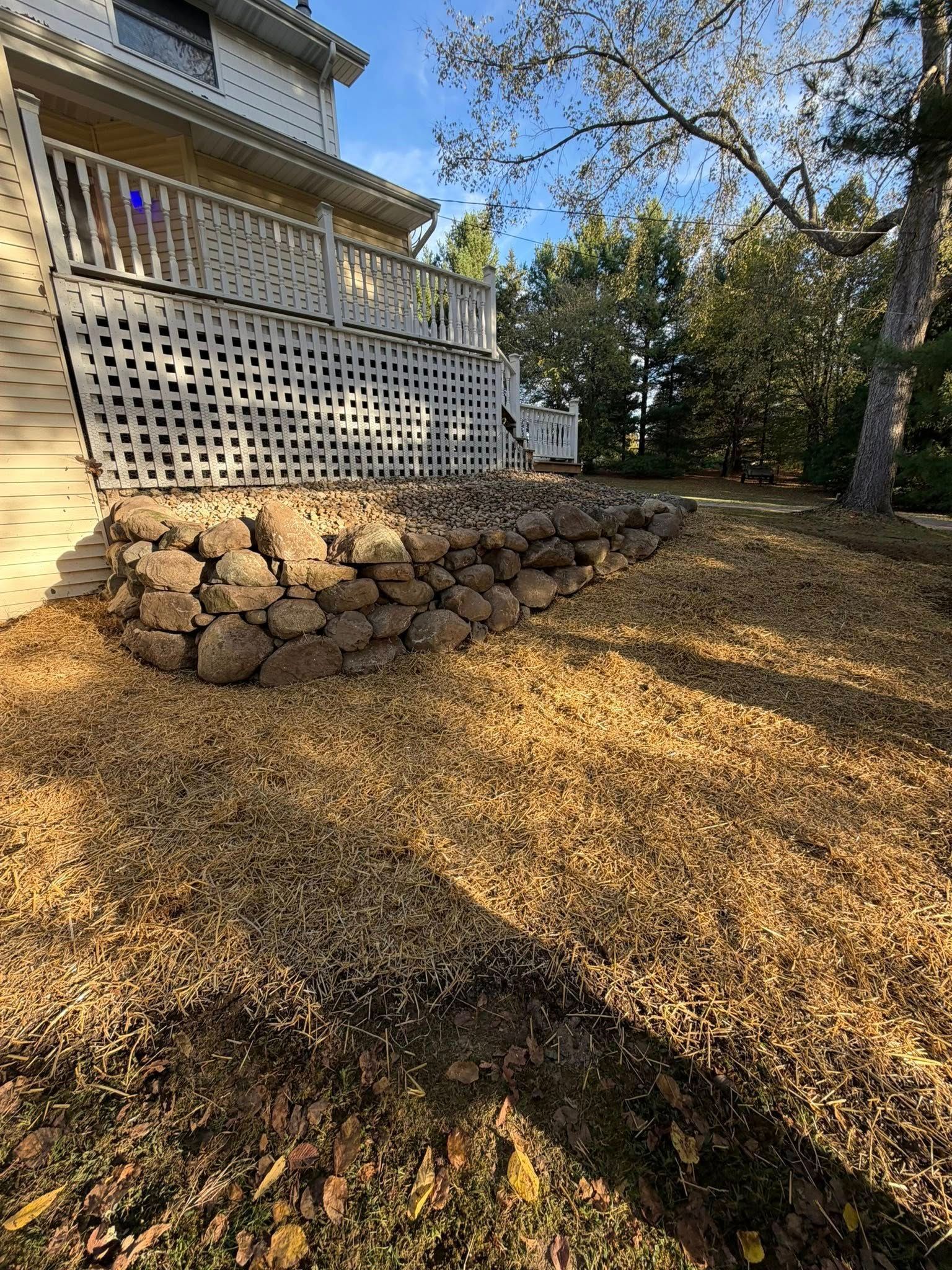 Stone retaining wall under a deck, surrounded by fallen leaves on a sunny day.