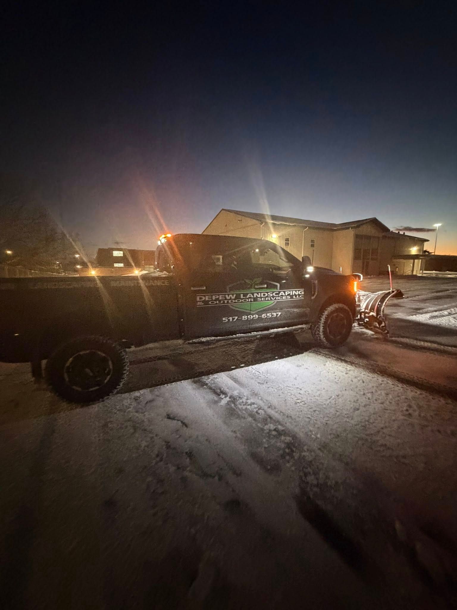 Dark truck with plow on snowy ground, lights on, building in background, at night.