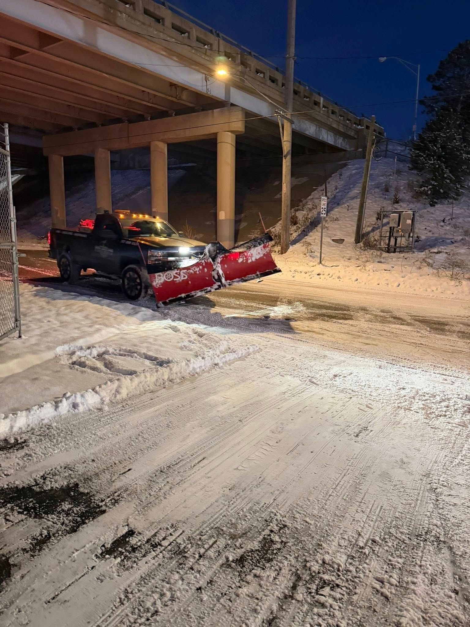 Snowplow truck under bridge clearing snow from road at dusk.