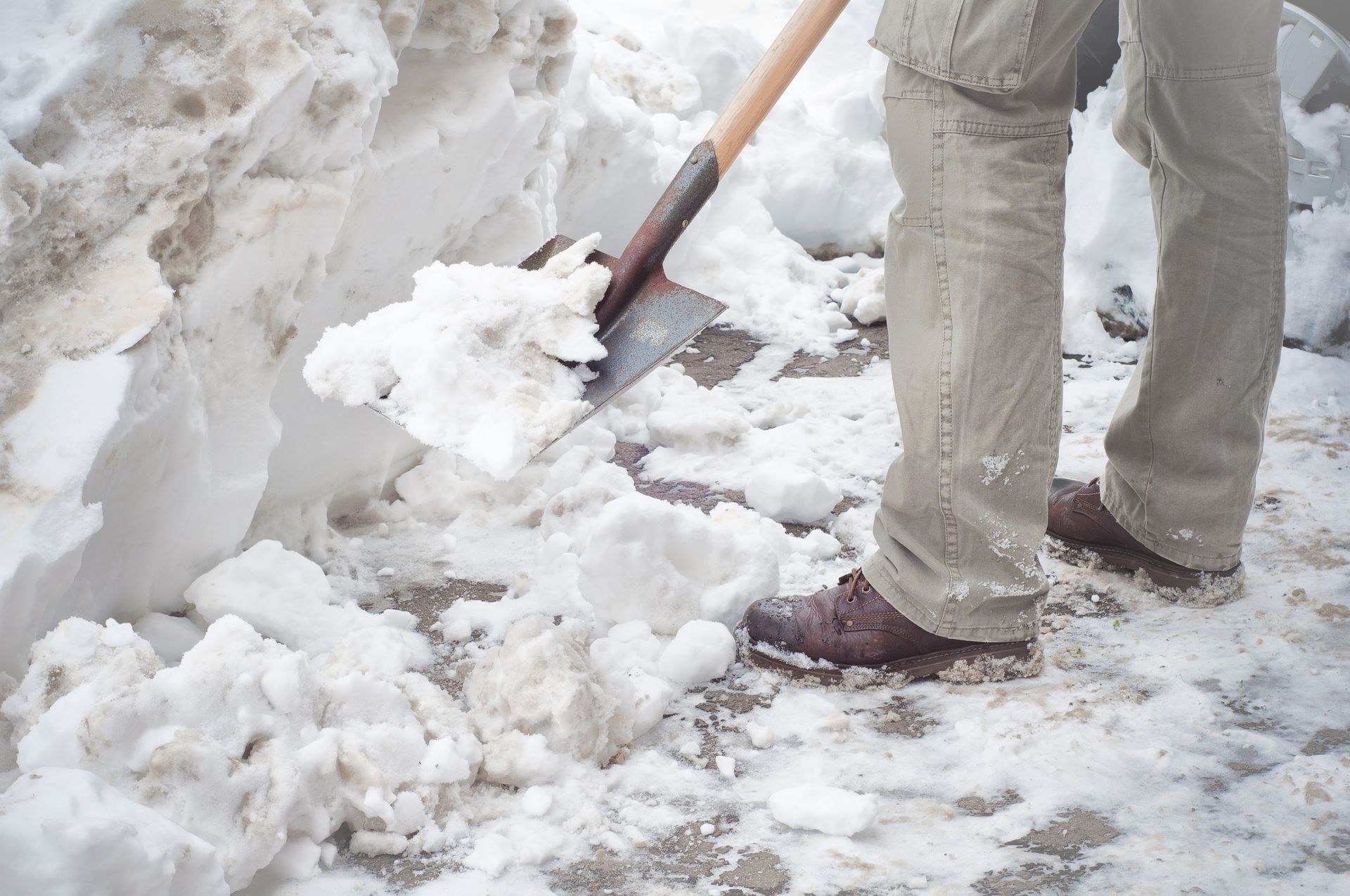 Person shoveling snow from a walkway, using a metal shovel. Brown shoes and khaki pants are visible.