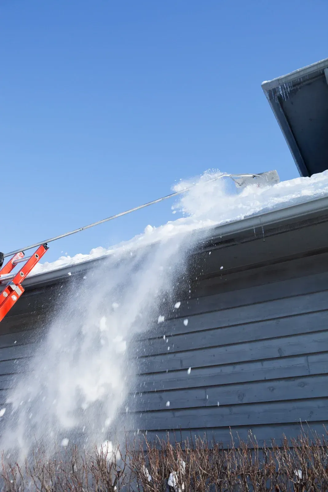 Snow being shoveled from a roof gutter with an extended snow rake under a blue sky.
