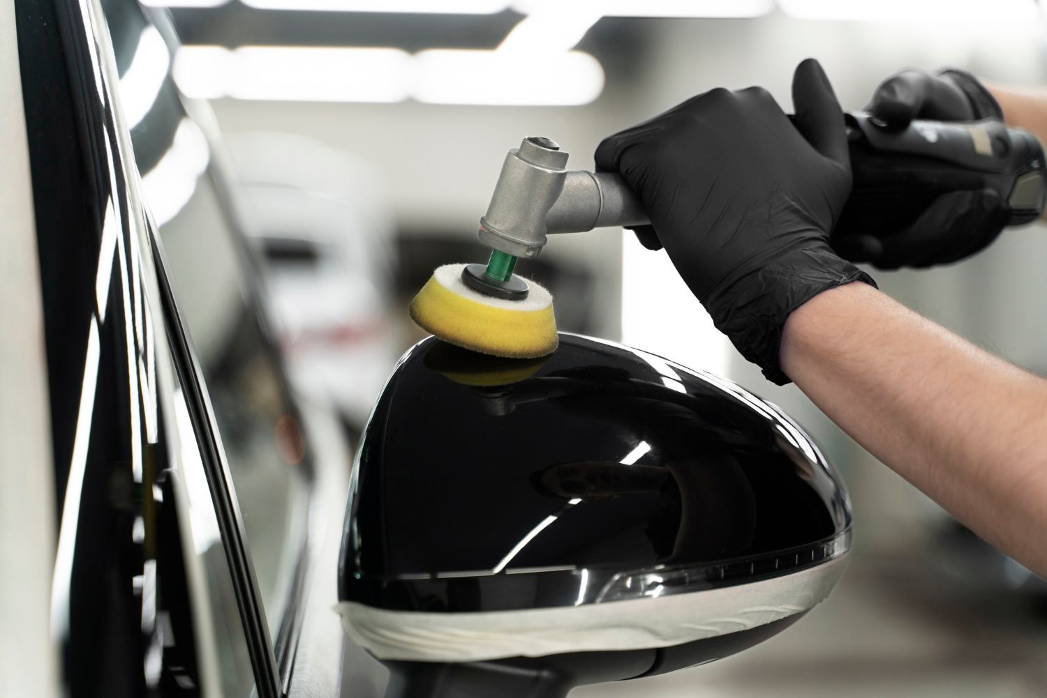 A person wearing black gloves uses a rotary polisher with a yellow pad on a glossy black car side mirror.