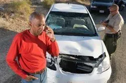 A man in a red shirt talks on a phone while an officer writes notes in front of a heavily damaged white car.