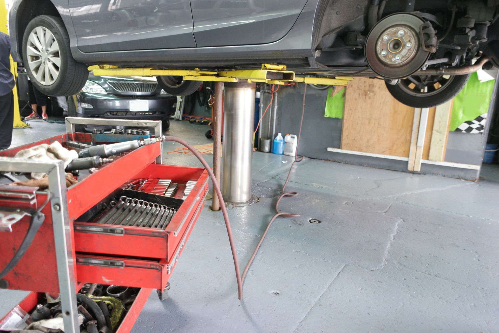 A grey sedan raised on an automotive lift in a workshop, with a red tool cart in the foreground.