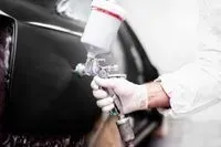 A person wearing white protective gloves uses a paint spray gun to apply a dark coating to a vehicle panel.