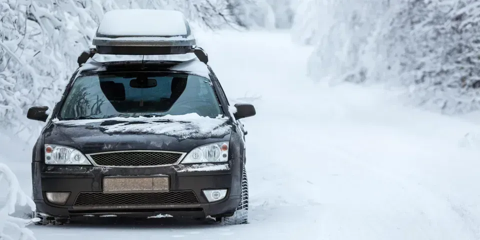 A black car with a roof cargo box drives down a snow-covered road flanked by trees.