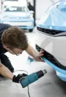 A person in a blue shirt kneels to mask a gray car in a workshop, using green paper and yellow tape.