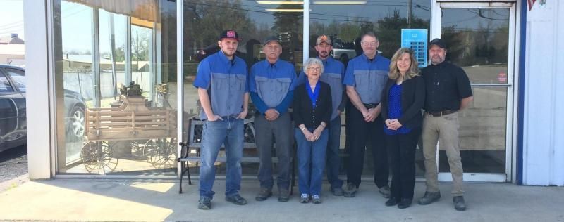 A group of people posing in front of an auto shop. Several are wearing blue and gray shirts.