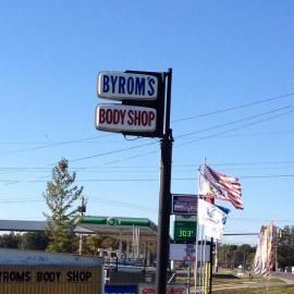 Sign for Byrom's Body Shop: blue sky, body shop sign, gas station, and American flag.