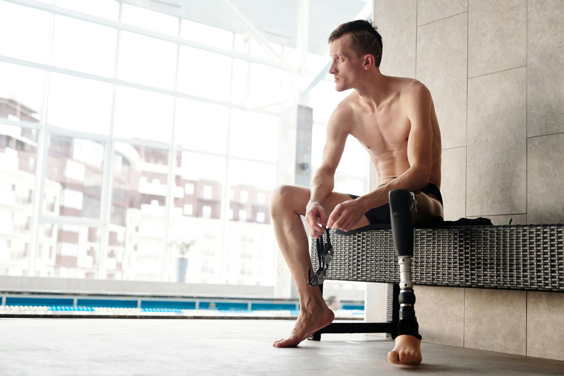 Man with a prosthetic leg sits by pool, looking out a large window.