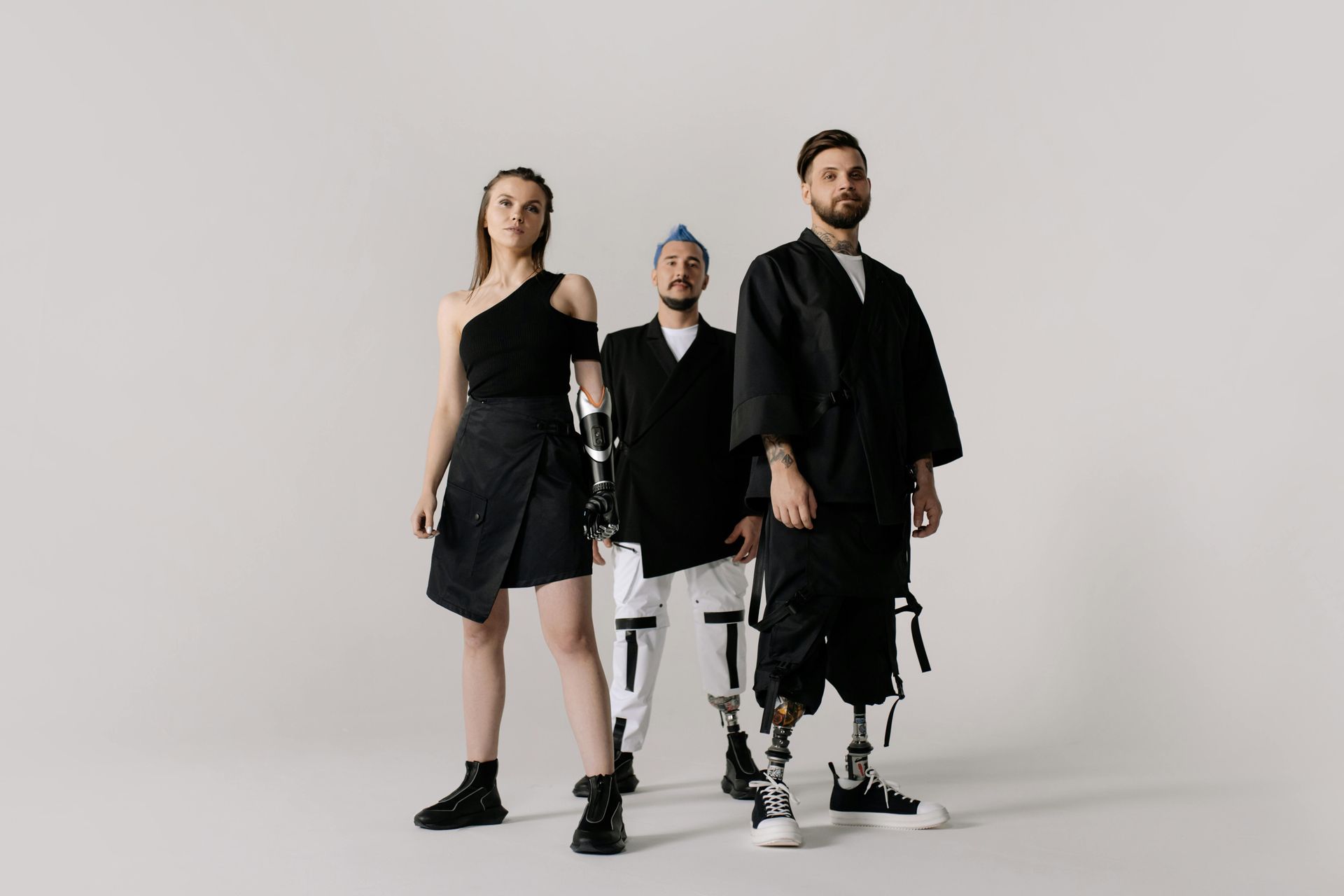 Three stylish people posing in a studio. Woman in black, man with blue hair, and man with prosthetic legs.