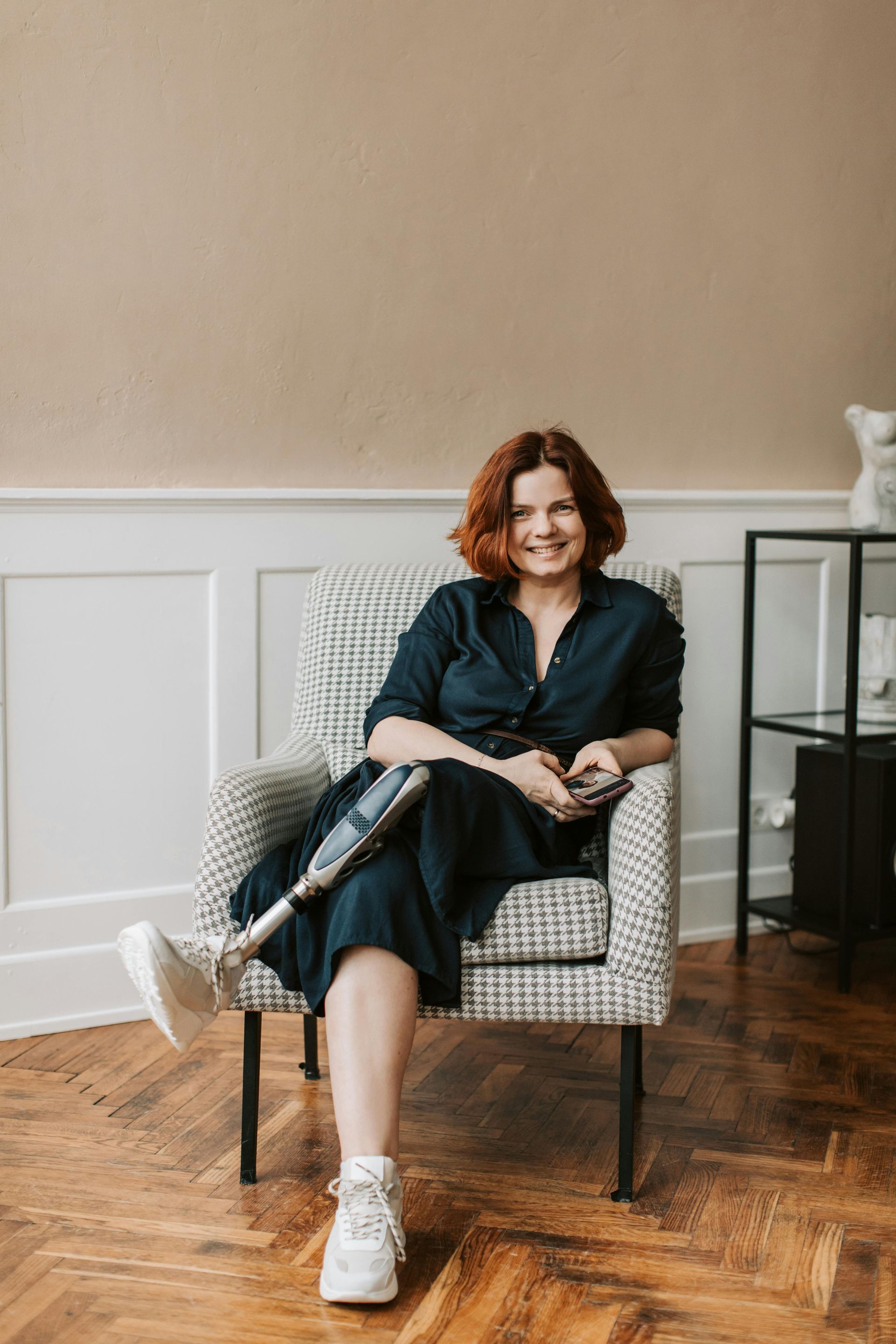 Woman with a prosthetic leg sits smiling in a patterned armchair. She wears a navy dress, white sneakers, and has red hair.