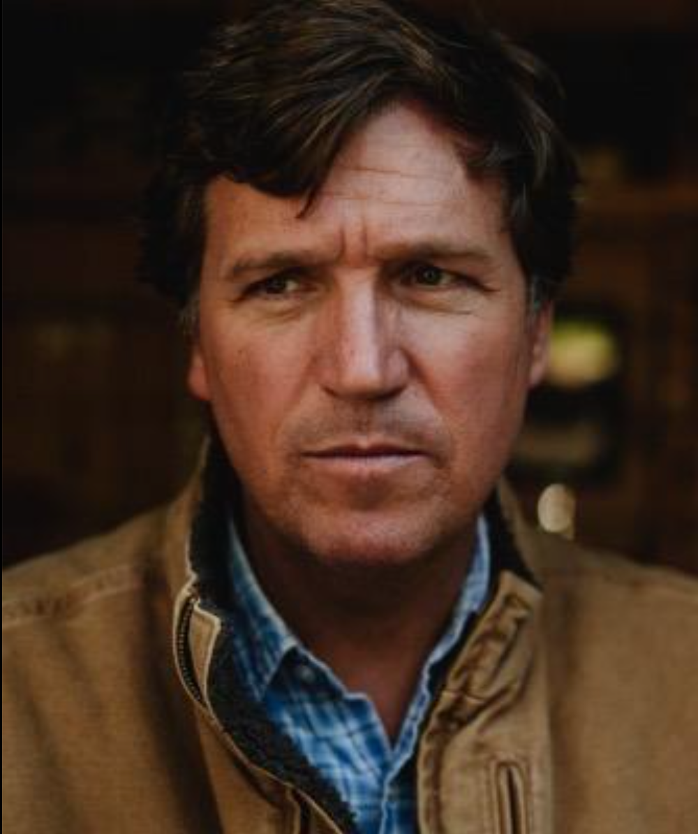 A man wearing a plaid shirt and a brown jacket looks at the camera