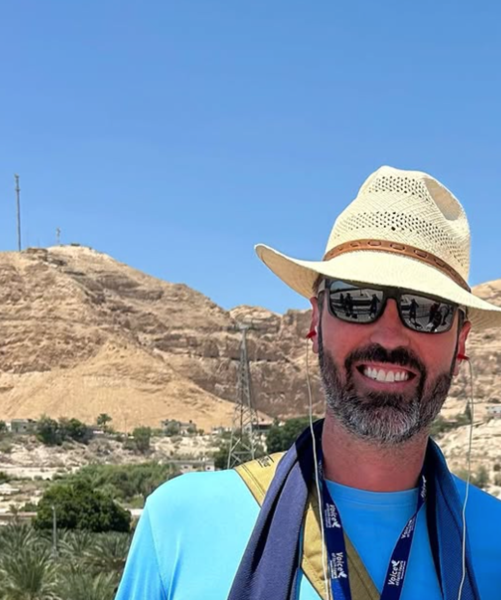A man wearing a hat and sunglasses stands in front of a mountain