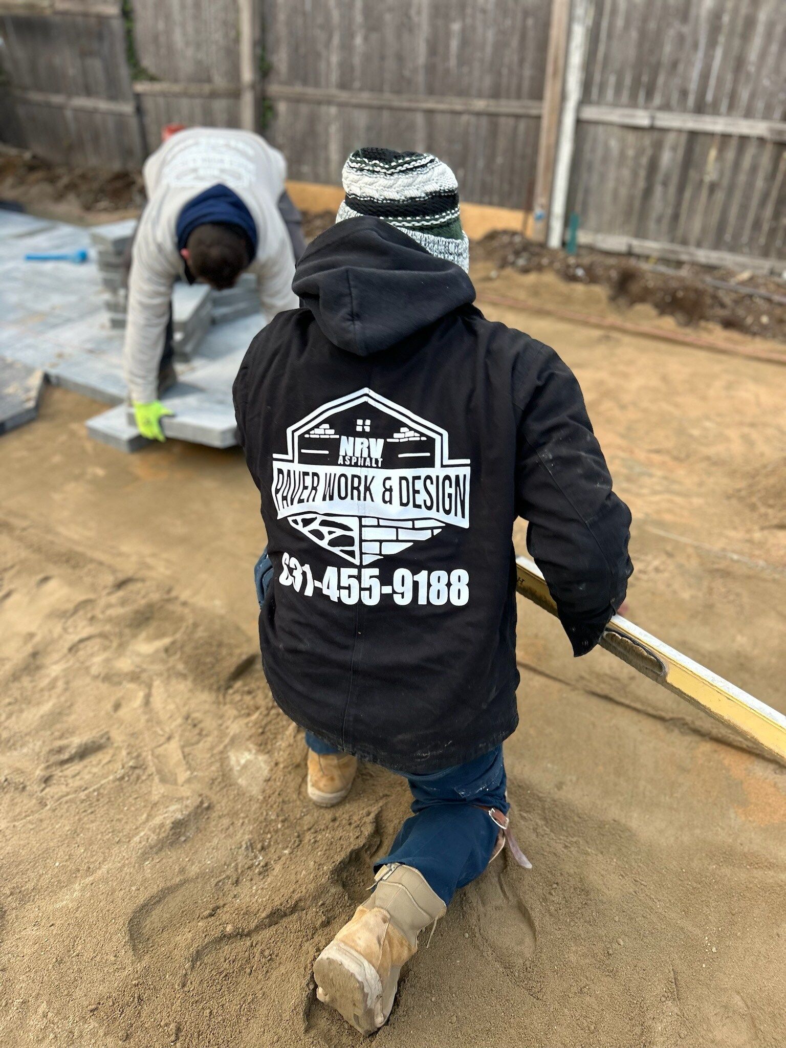A person wearing a black jacket with a logo on the back is kneeling in the sand.
