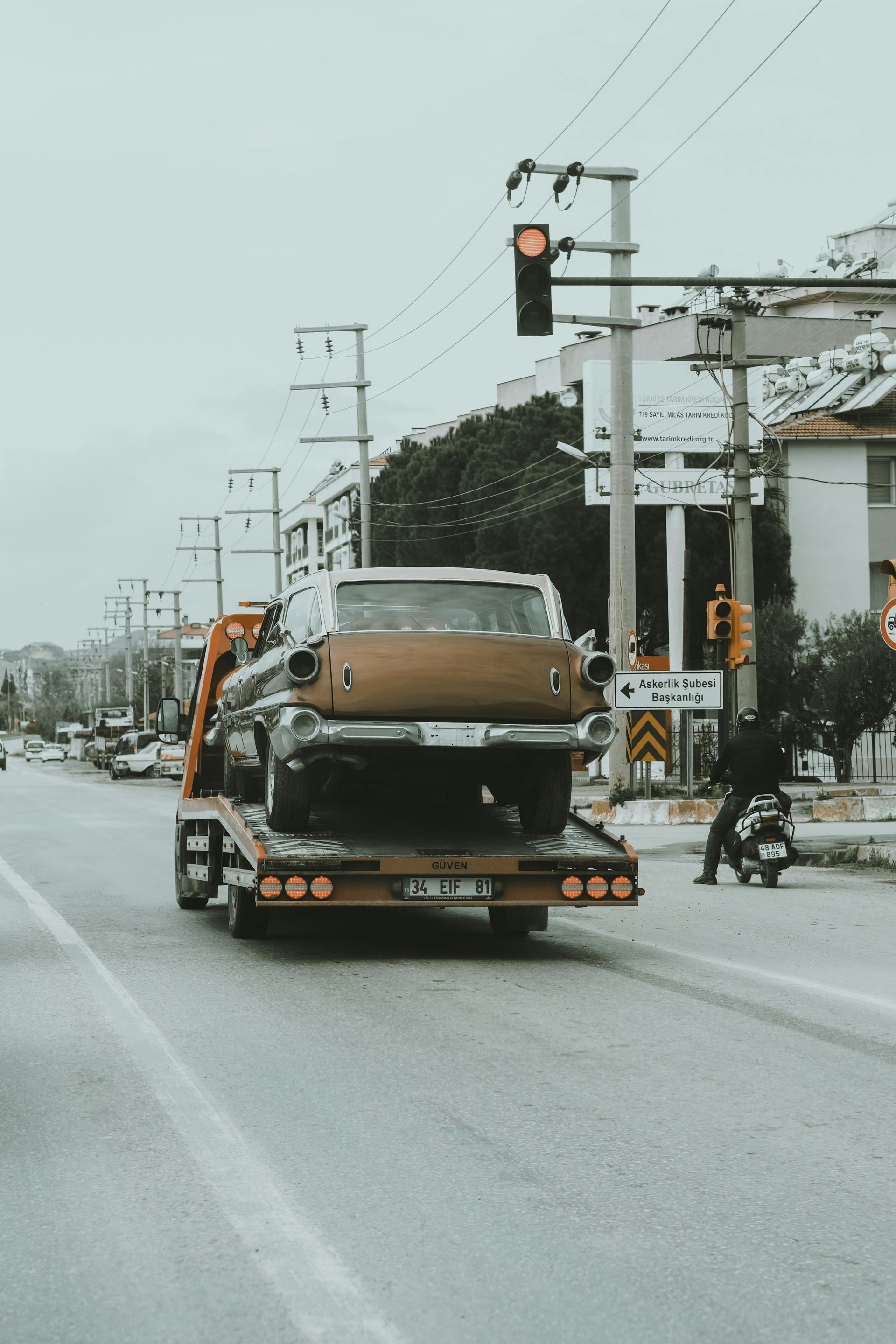 Tow truck carrying a classic car on a city street under a cloudy sky.