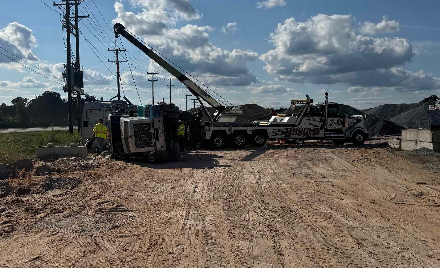 Tow truck lifting a toppled white truck from a muddy roadside near power lines on a cloudy day.