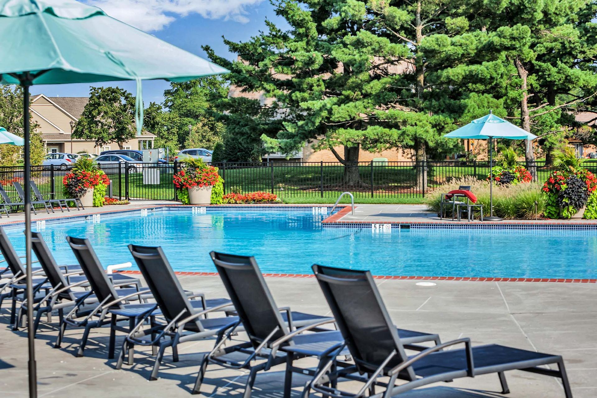 Outdoor apartment pool with blue water, lounge chairs, teal umbrellas, and landscaped surroundings.