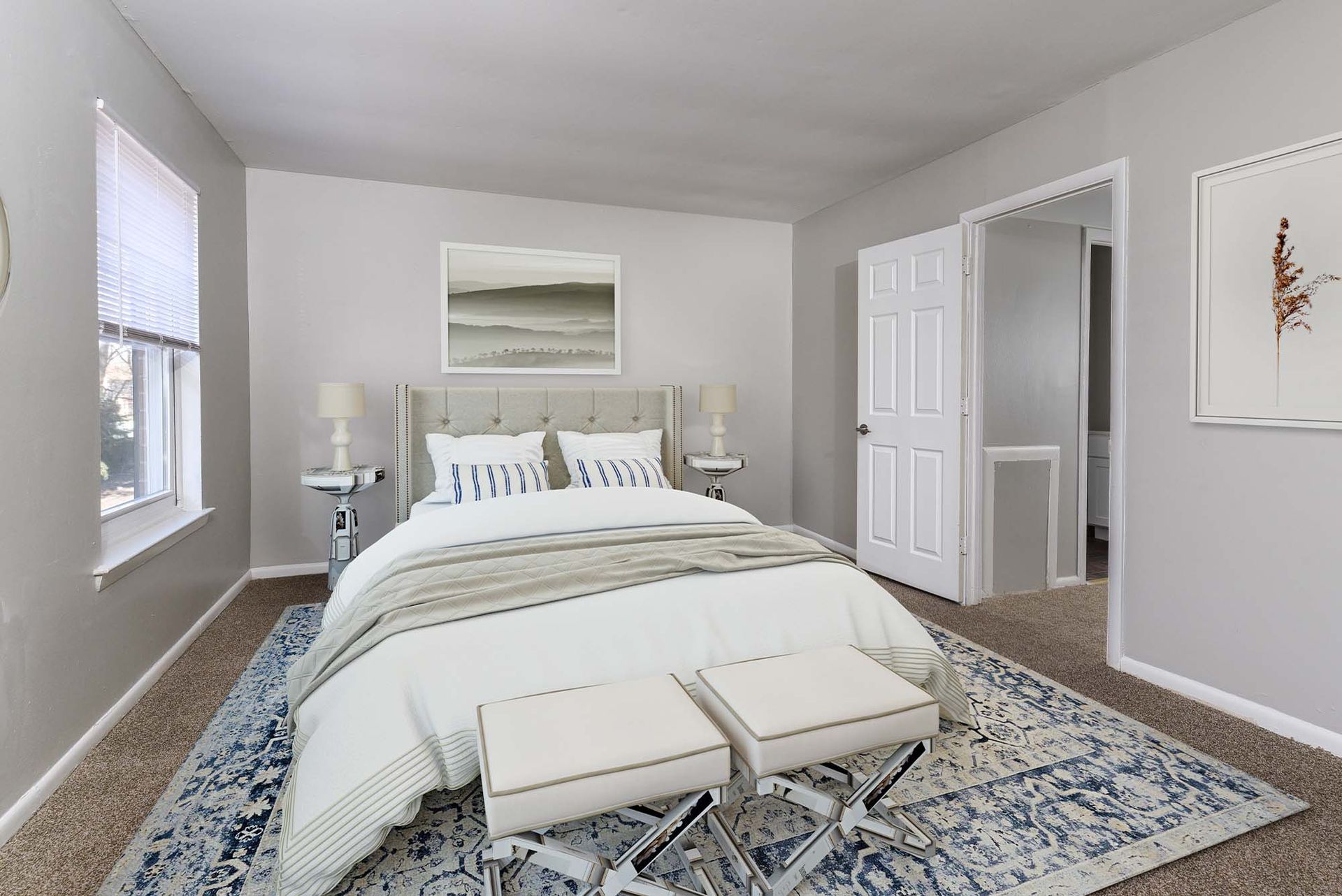 Bedroom with tufted headboard, two nightstands, lamps, white bedding, and two ottoman benches on a blue rug.
