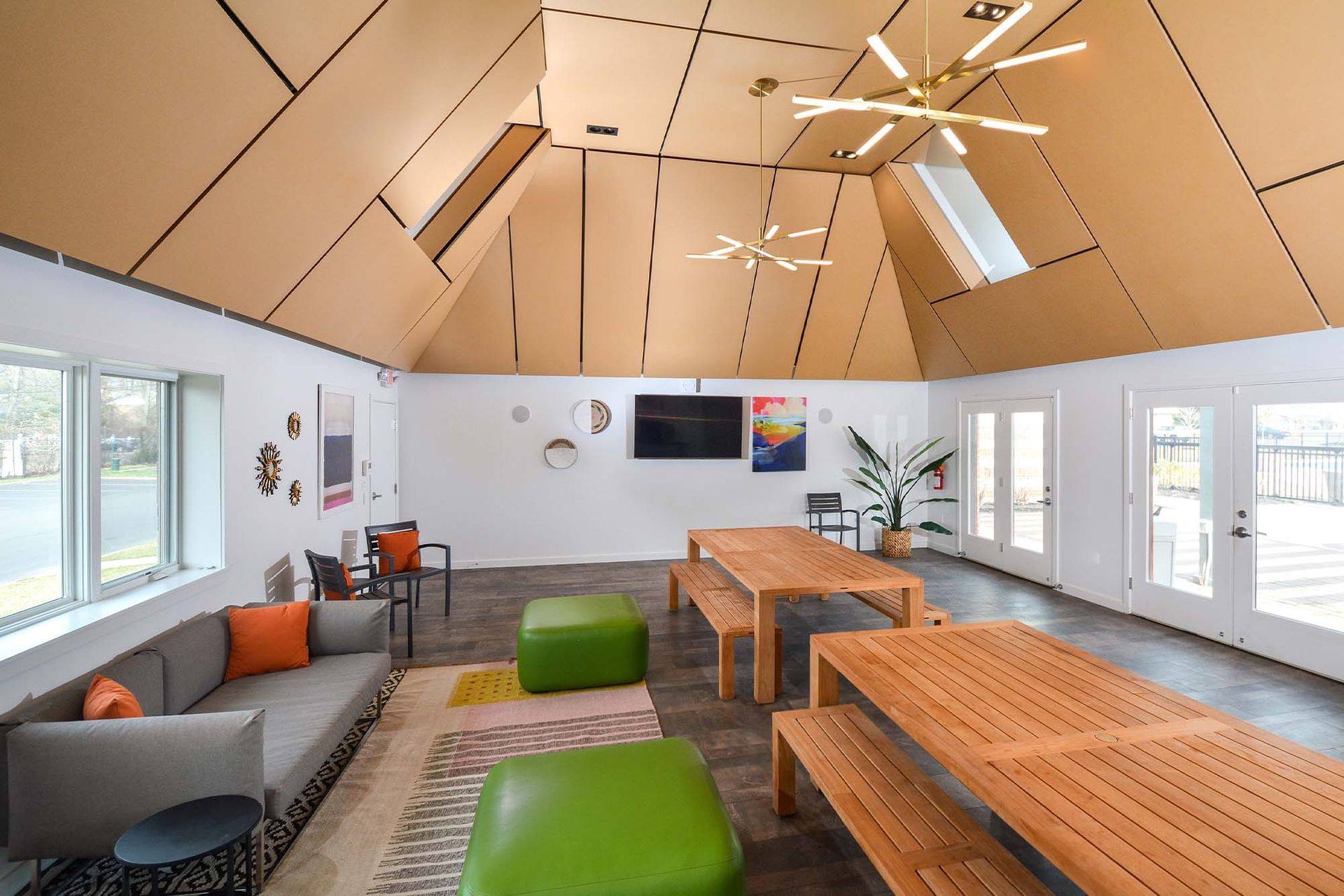 Bright community lounge with long wooden tables, cushioned seating, and a wall-mounted TV.