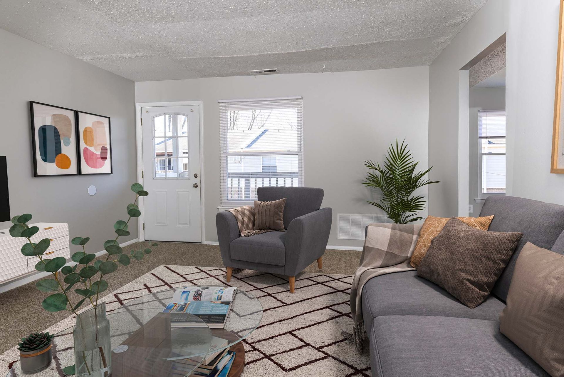 Living room in an apartment with a gray sofa, armchair, glass coffee table, plants, and abstract wall art.