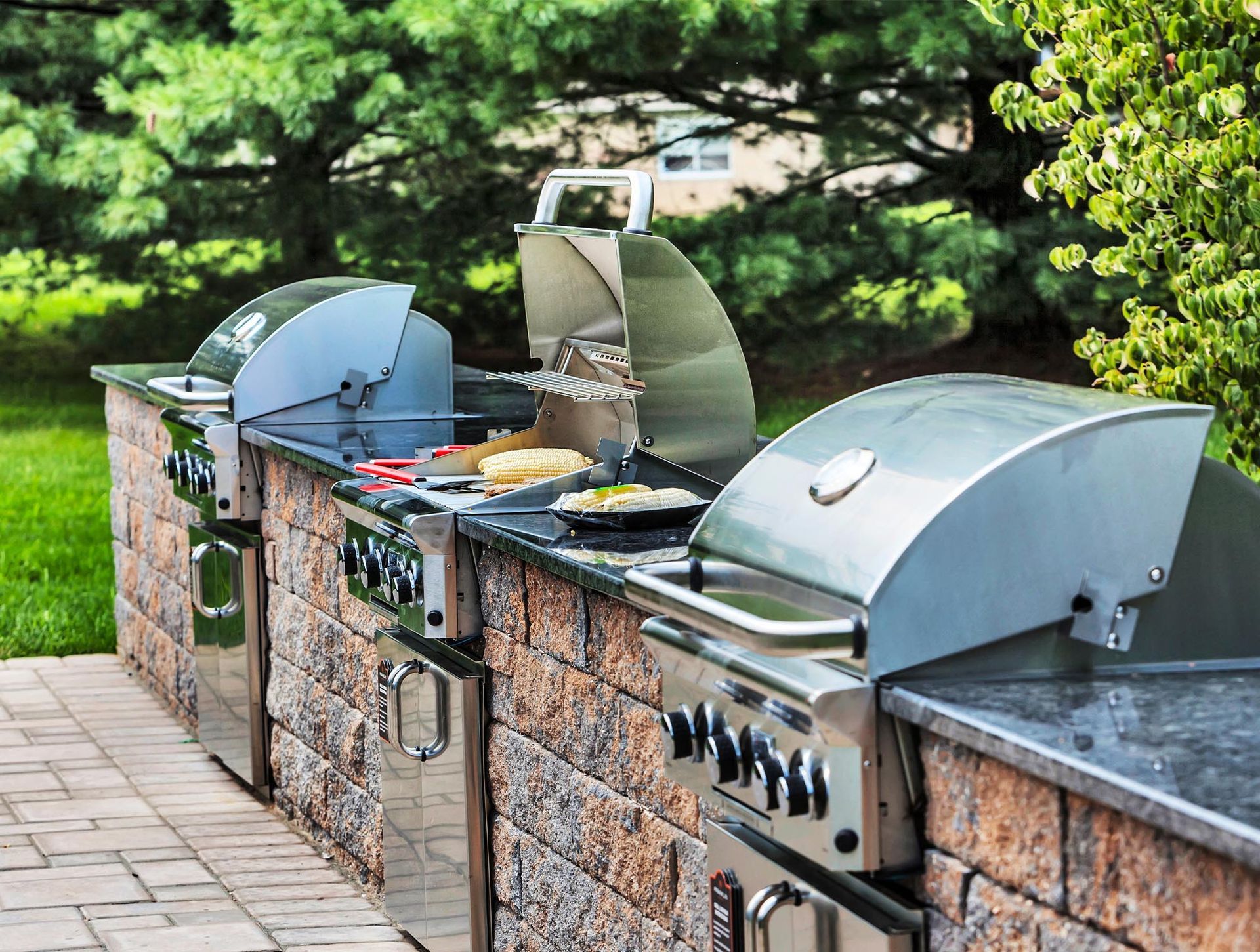 Outdoor stainless-steel built-in grills along a stone counter in a landscaped courtyard.