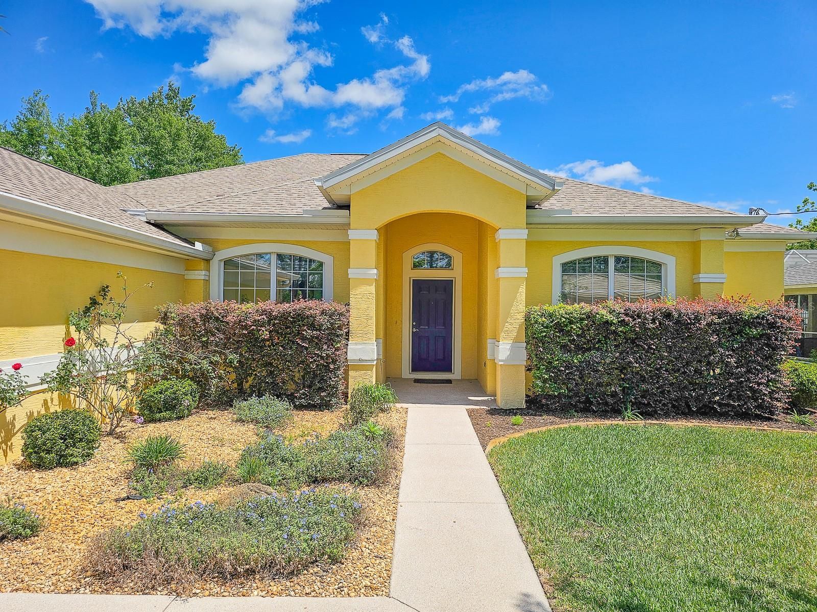 Yellow house with purple front door, windows, and neatly trimmed bushes in front.