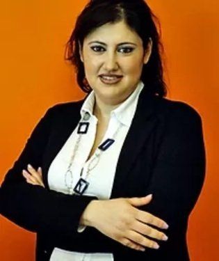 Donna in blazer nero, braccia incrociate, sorridente. Indossa una camicia bianca e una collana su sfondo arancione.