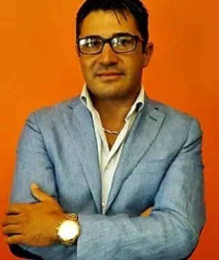 Uomo con giacca blu, occhiali e orologio, braccia incrociate su sfondo arancione.