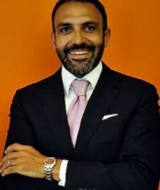 Uomo in giacca e cravatta, braccia incrociate, sorridente, con cravatta rosa e orologio, su sfondo arancione.