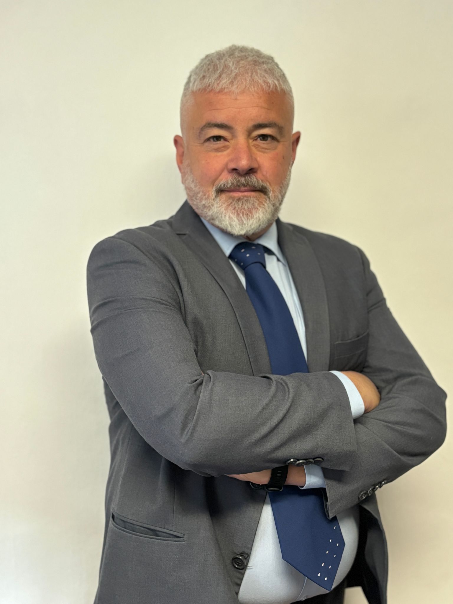 Uomo in giacca e cravatta con le braccia incrociate, in piedi davanti a uno sfondo bianco.