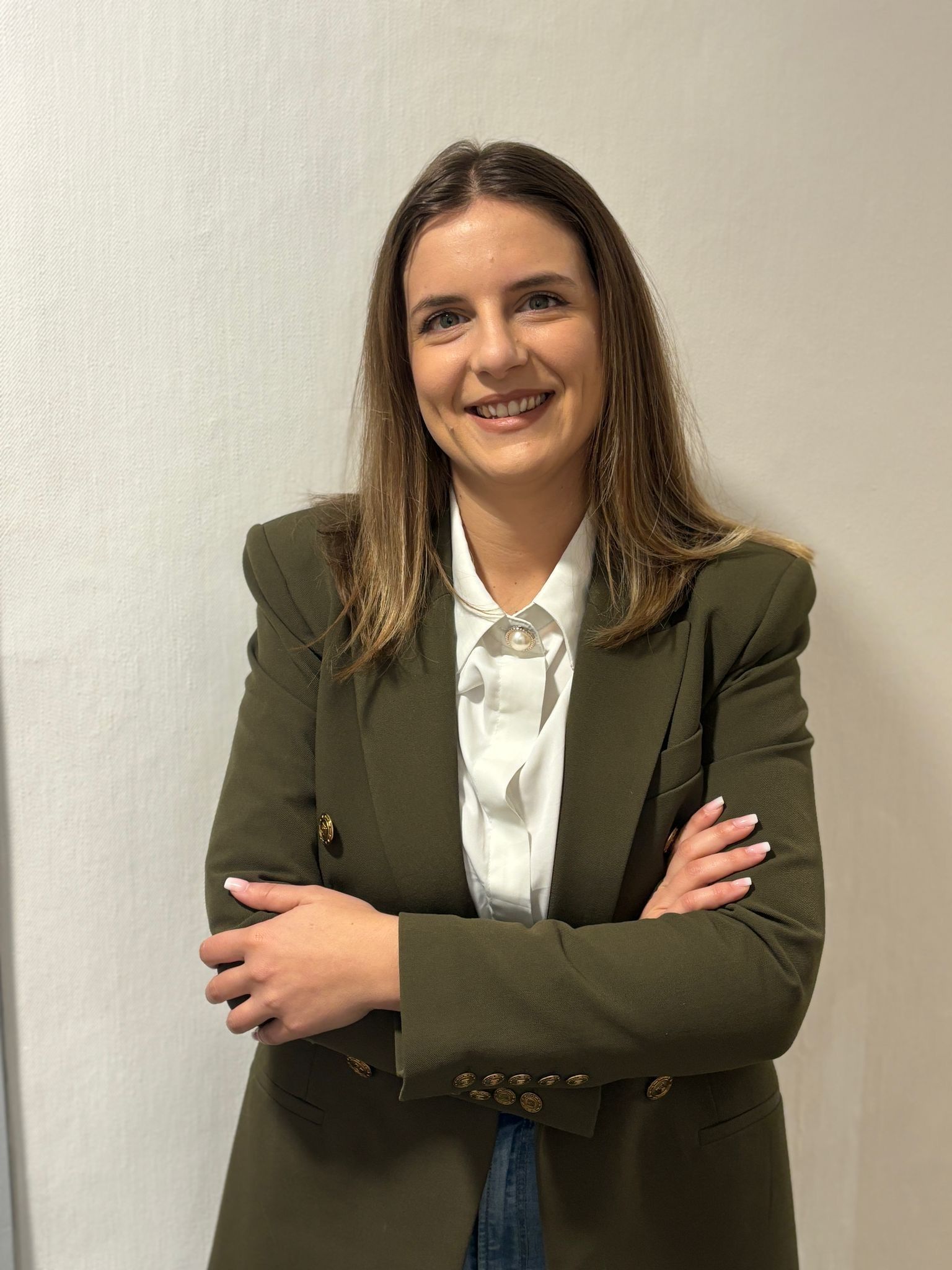 Donna con giacca verde oliva e camicia bianca, braccia incrociate, sorridente.