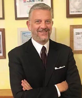 Uomo in abito scuro, con le braccia incrociate, sorridente, davanti a documenti incorniciati.