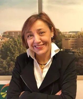 Una donna in giacca e camicia bianca sorride, con le braccia incrociate, in ufficio, mentre guarda la telecamera.