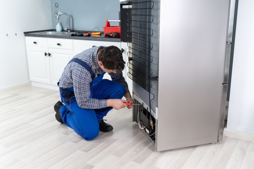 Technician repairs refrigerator in a kitchen, kneeling and using a screwdriver.