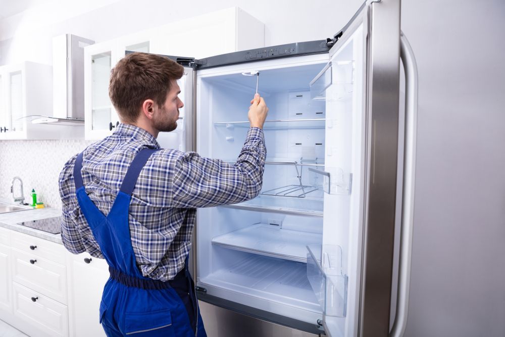 Man in blue overalls inspecting the inside of a refrigerator with an open door in a kitchen.
