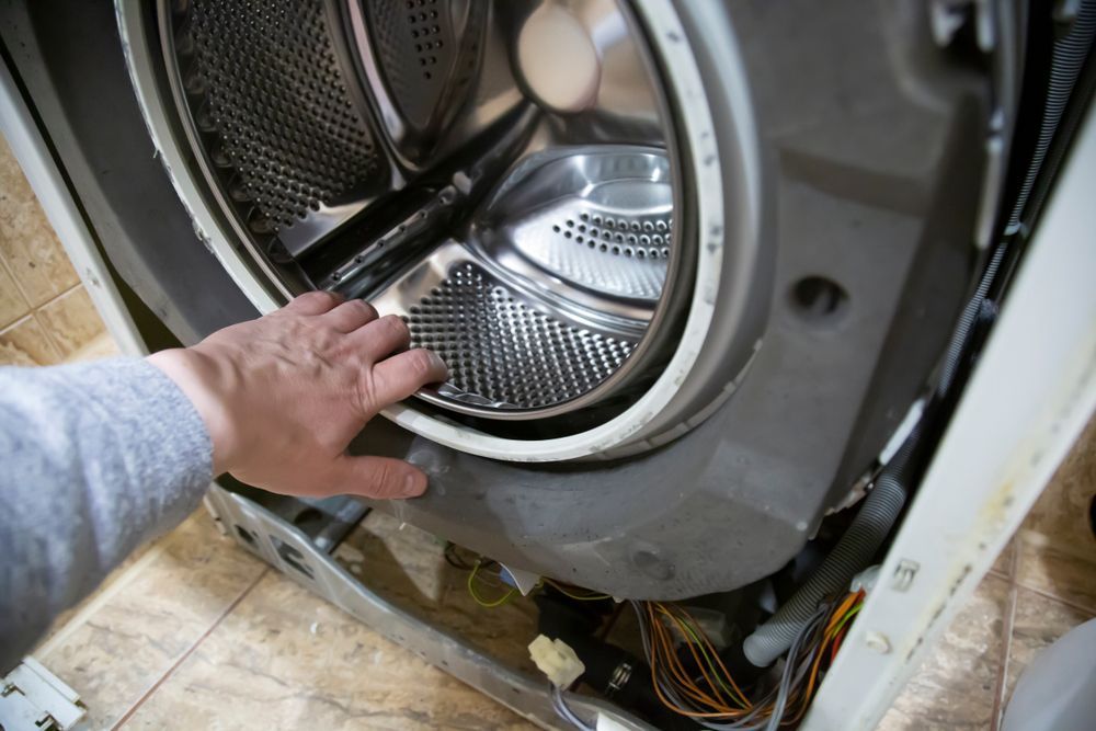 Hand touching a washing machine's rubber seal, open view of the drum, inside a tiled room.