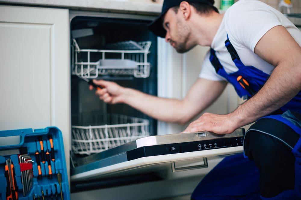 Technician in blue overalls repairing a dishwasher with tools, kneeling in front of the appliance.