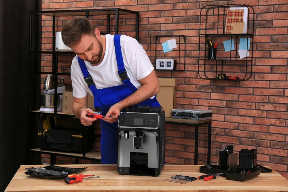 A man in blue overalls repairs a coffee machine with a screwdriver on a table near a brick wall.