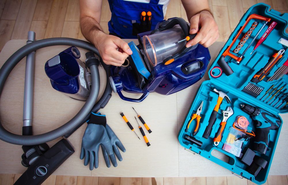 Person repairing a blue vacuum cleaner with tools, including a toolbox and screwdriver on a table.
